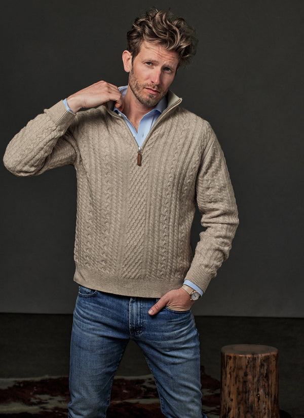 Man wearing a beige cable knit sweater and blue jeans against a dark background