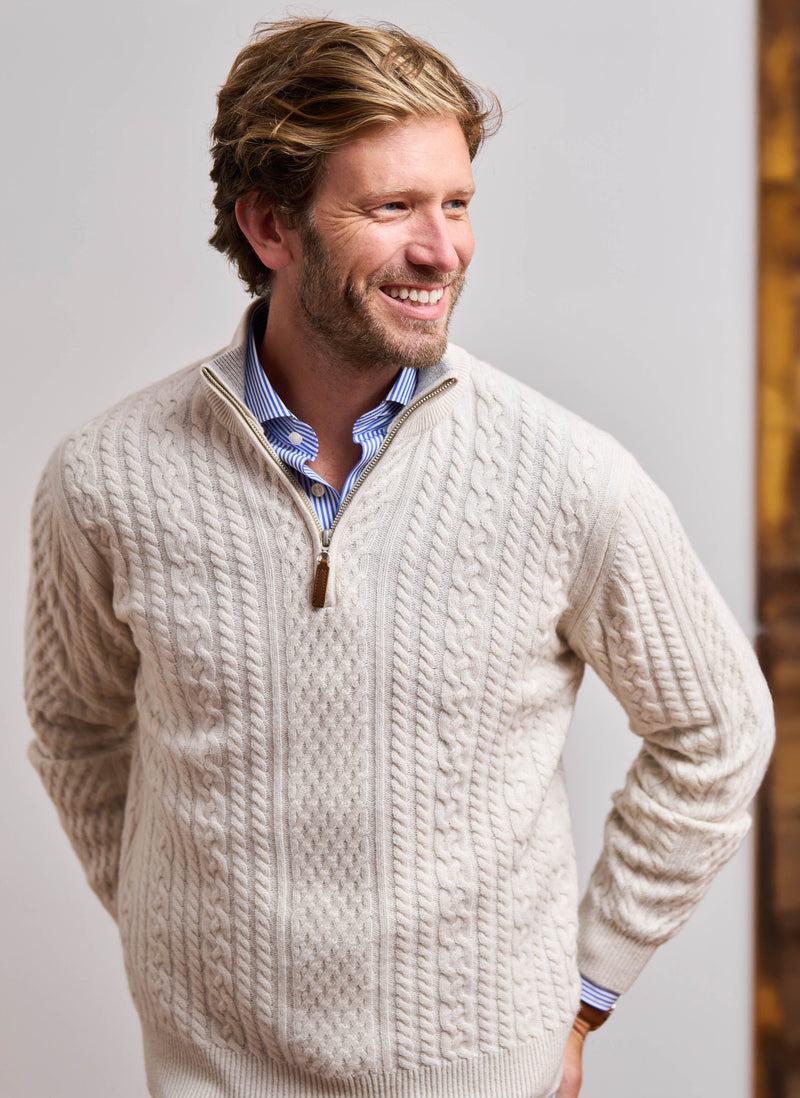 Man wearing a beige cable knit sweater over a blue shirt against a neutral background