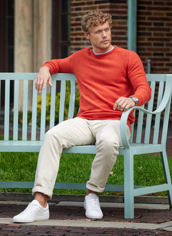model sitting on a light blue bench with a light grey undershirt, 100% merino wool crew neck sweater in rust mélange, khaki pants, and white sneakers