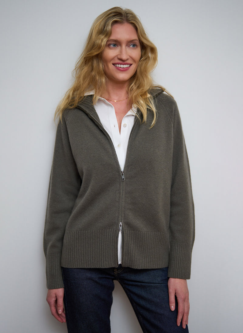 Woman wearing a green zip-up hoodie over a white shirt against a plain background