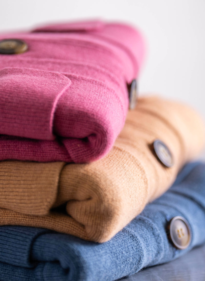 Stack of folded sweaters in pink, beige, and blue with buttons on a light background