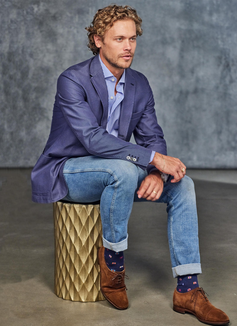 model wearing a blue stripe shirt with navy blue sports coat sitting on a gold stool with cotton square socks in navy and brown suede shoes 